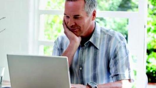 middle-aged man sitting at a computer with hand cupping chin, smiling